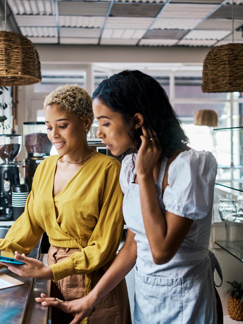 two ladies behind a coffee shop counter looking at paper work on a clip board and phone