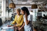 two ladies behind a coffee shop counter looking at paper work on a clip board and phone